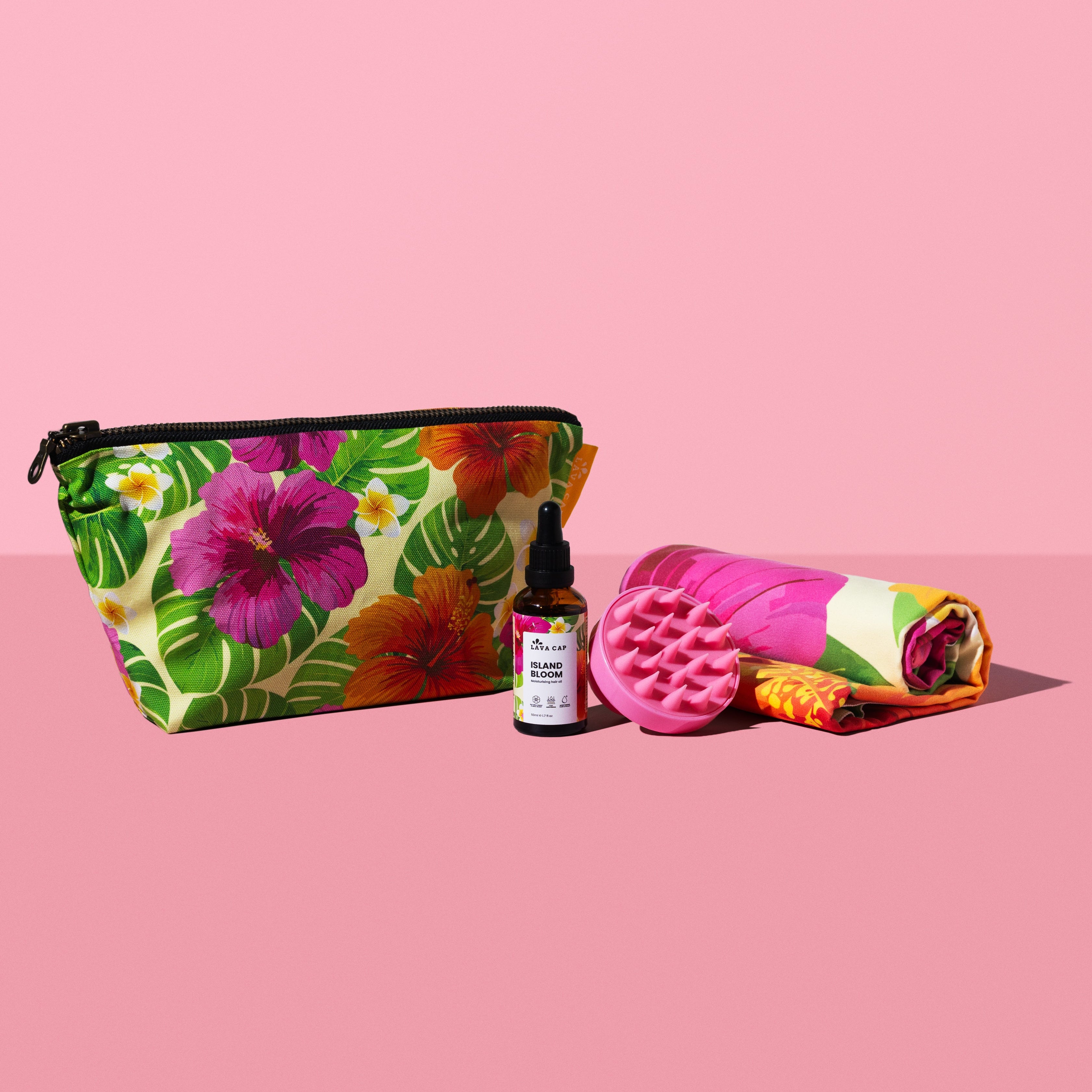 Floral toiletry bag, hair oil bottle, rolled-up matching floral hair towel and a pink scalp massager on a pink background