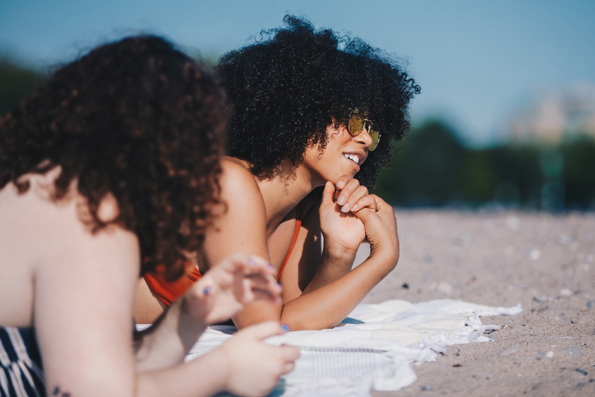 Two women with curly hair & sunglasses lying on a beach | Lava Cap microwavable conditioning cap nourishes curly hair that's prone to dryness